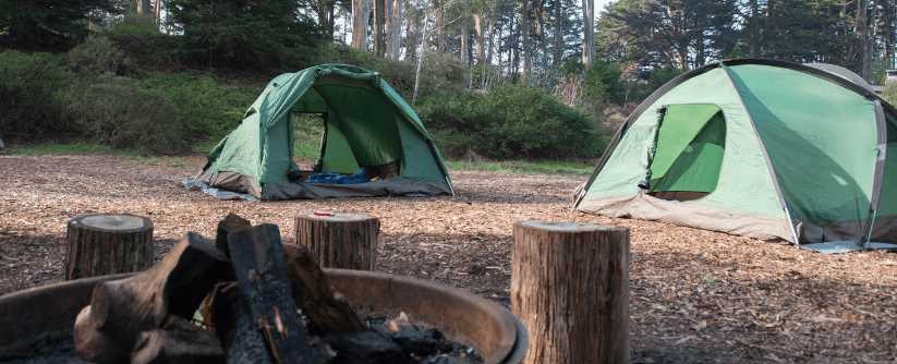 Two green tents sit in a campsite near a firepit, surrounded by trees.