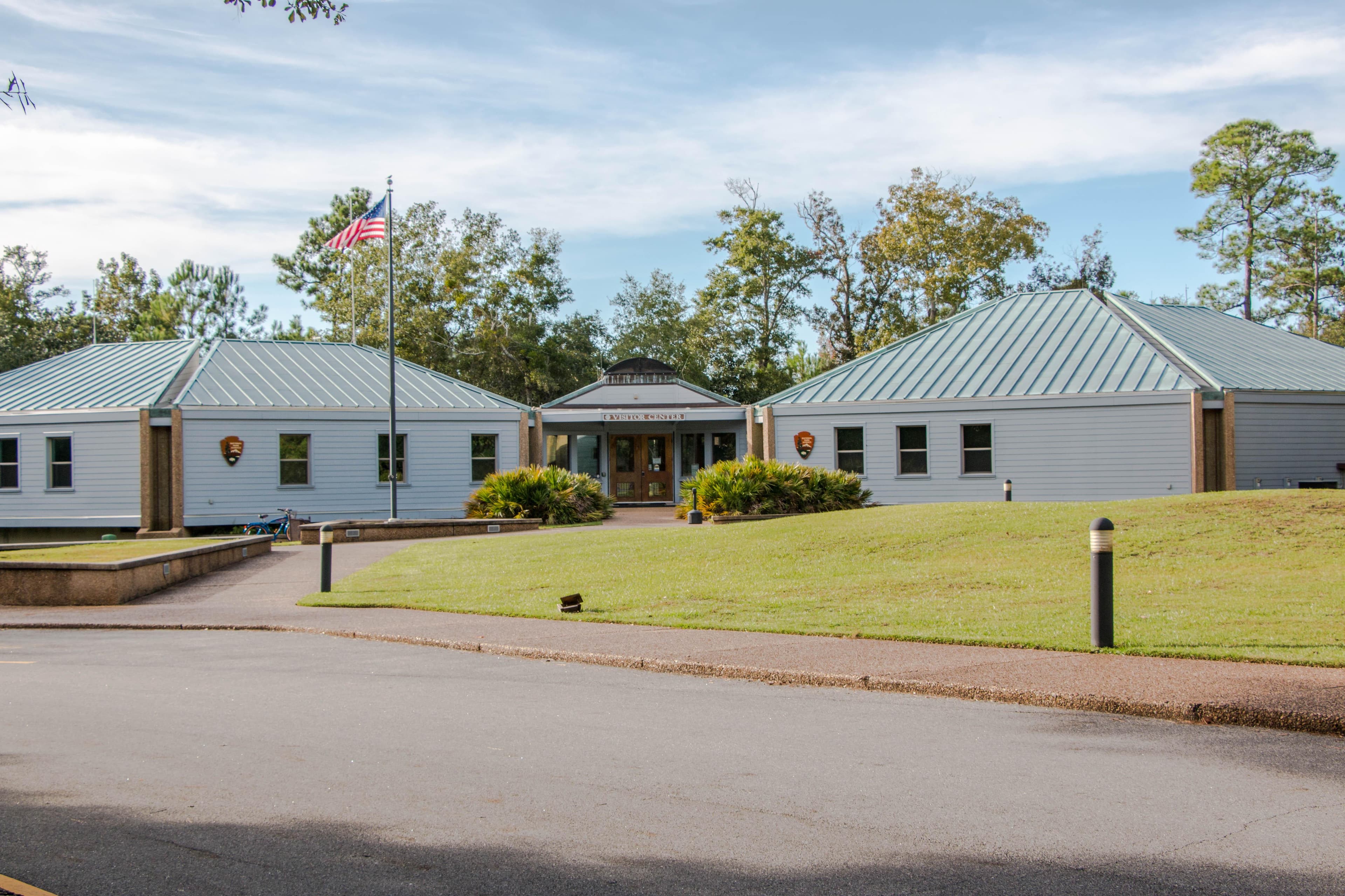 Grey buildings with a blue roof. Green grass and a road in the foreground.
