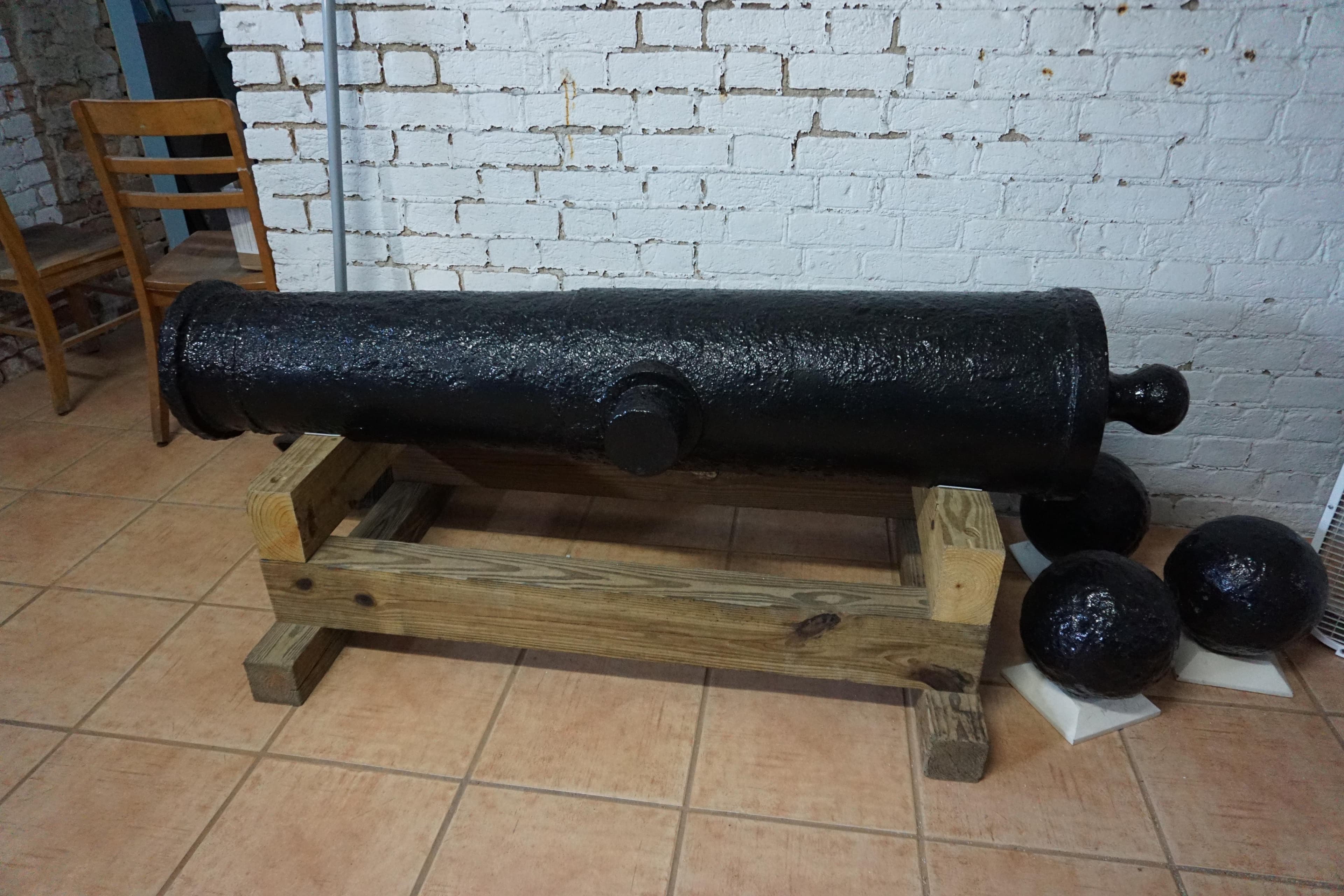 A preserved black cannon sits in a brown wooden frame in front of a white brick wall