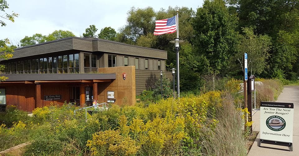 An American flag waves in the wind on a flag pole. A brown building nests within green vegetation.