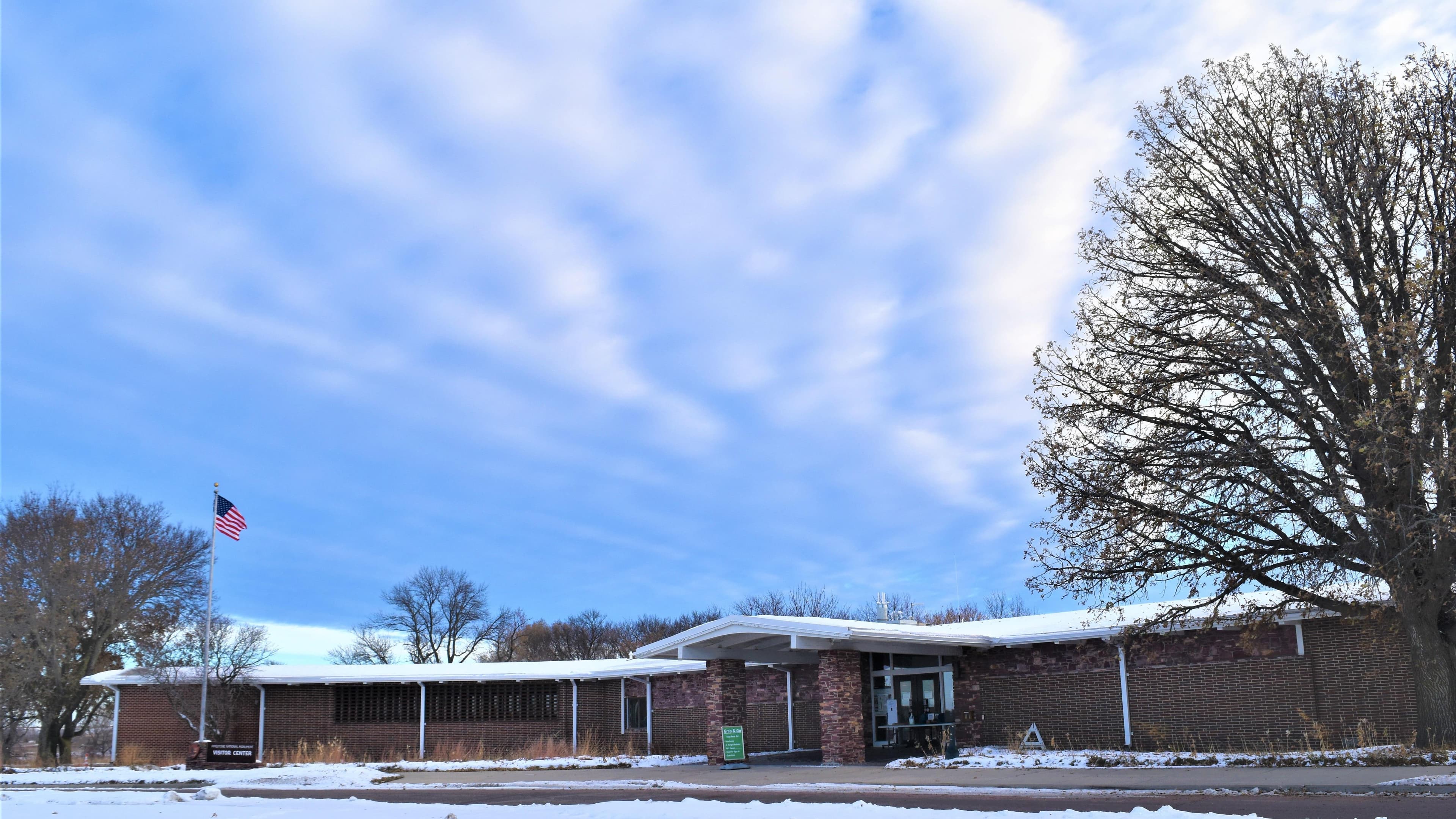 A low-profile, Mission 66-sytle building with a tree to the right, a US flag to the left, and clouds