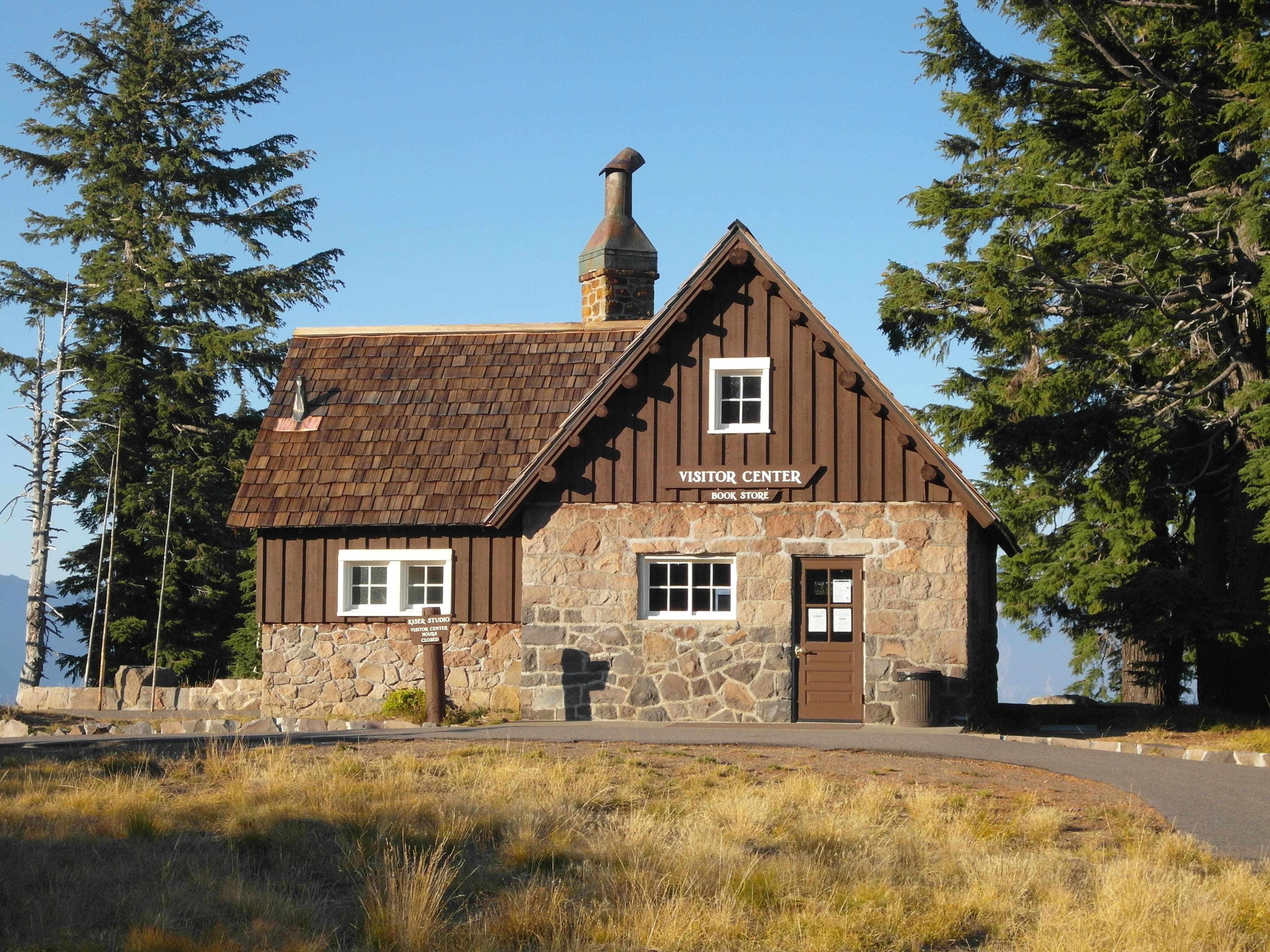 a native stone and wood building, sidewalks through a grassy meadow, towering conifers, and blue sky