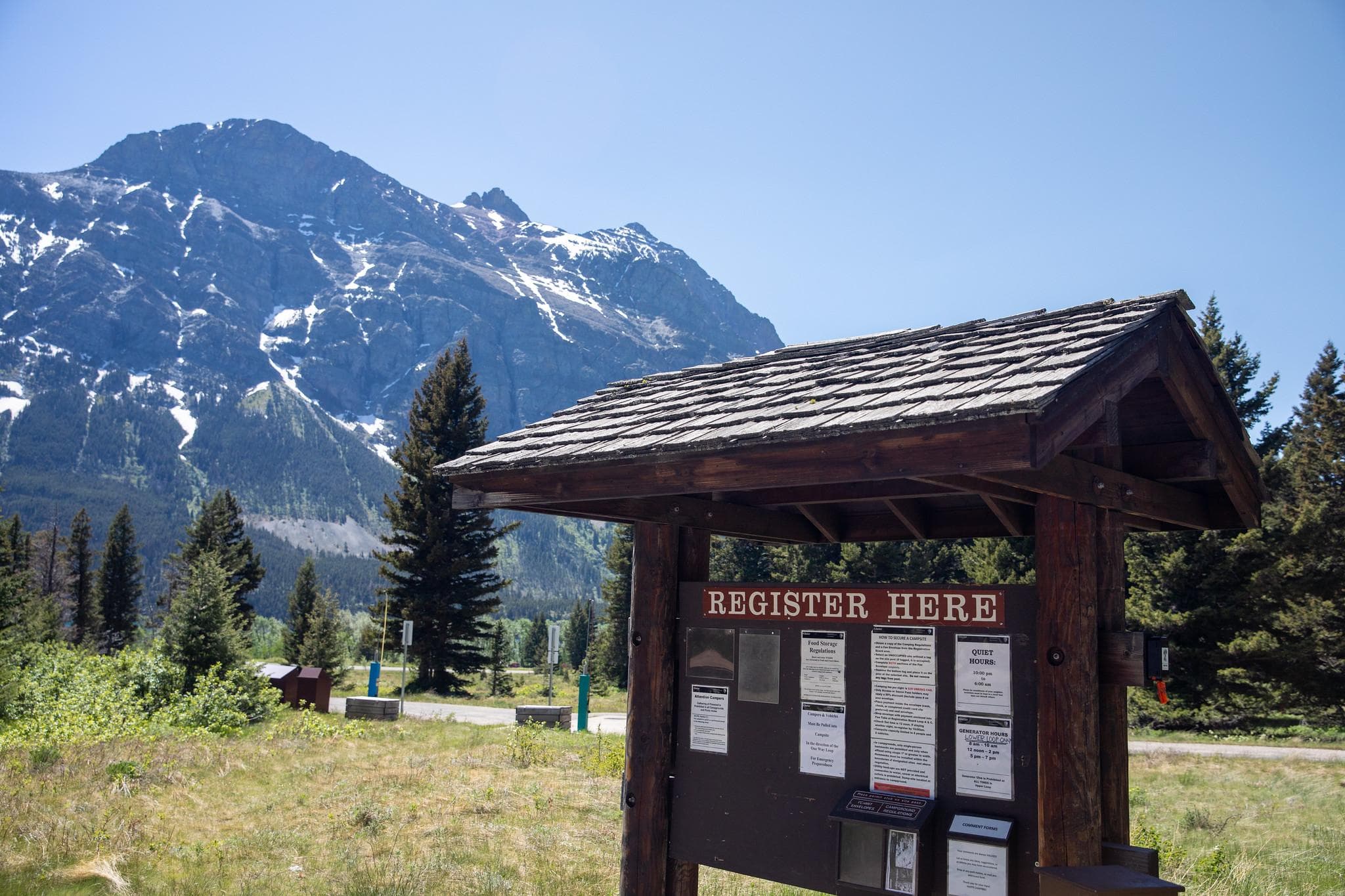 A small, wooden kiosk with a brown "Register Here" sign stands in front of a large mountain.