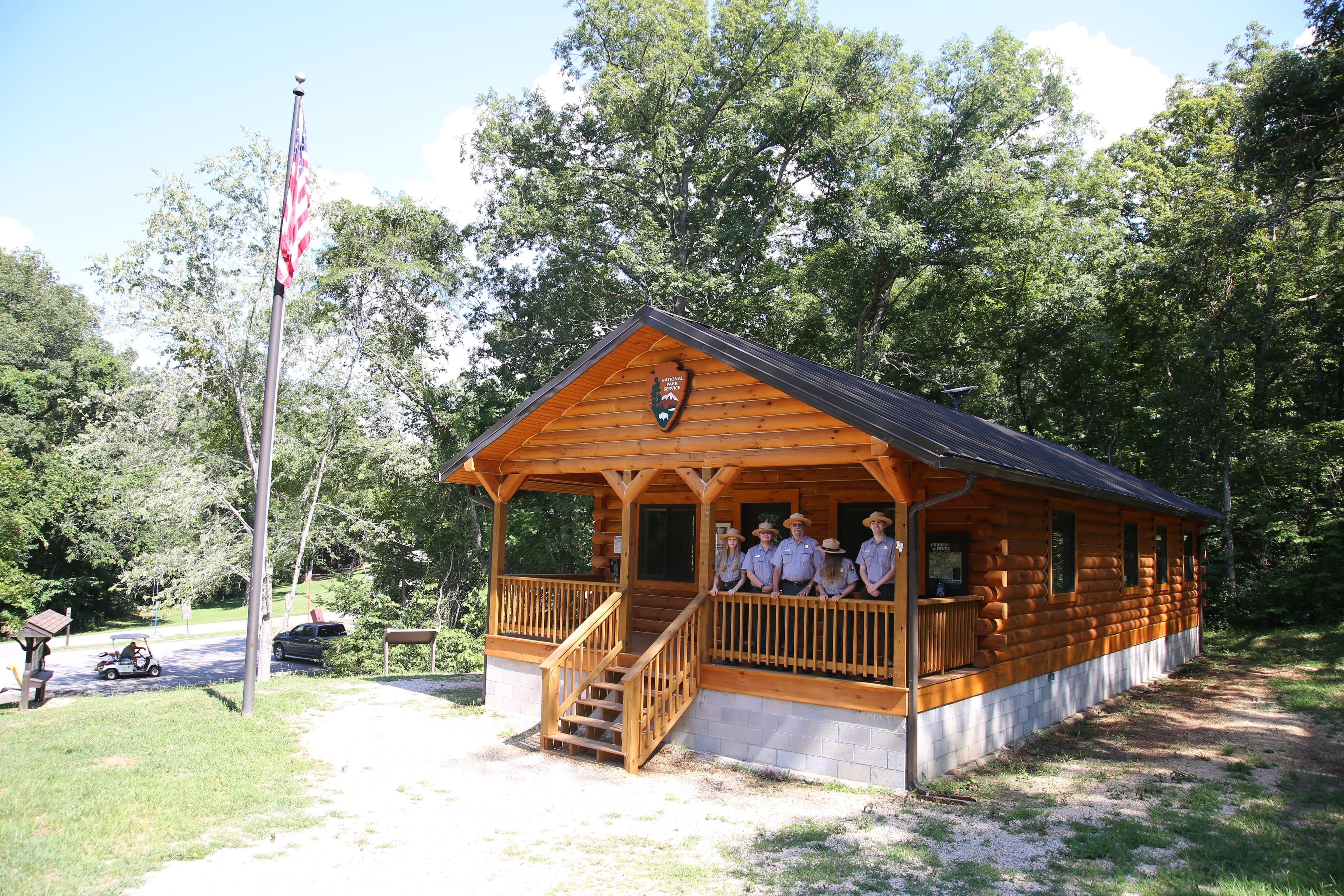 A log building sits atop a grassy hillside. Five rangers stand on the front porch.