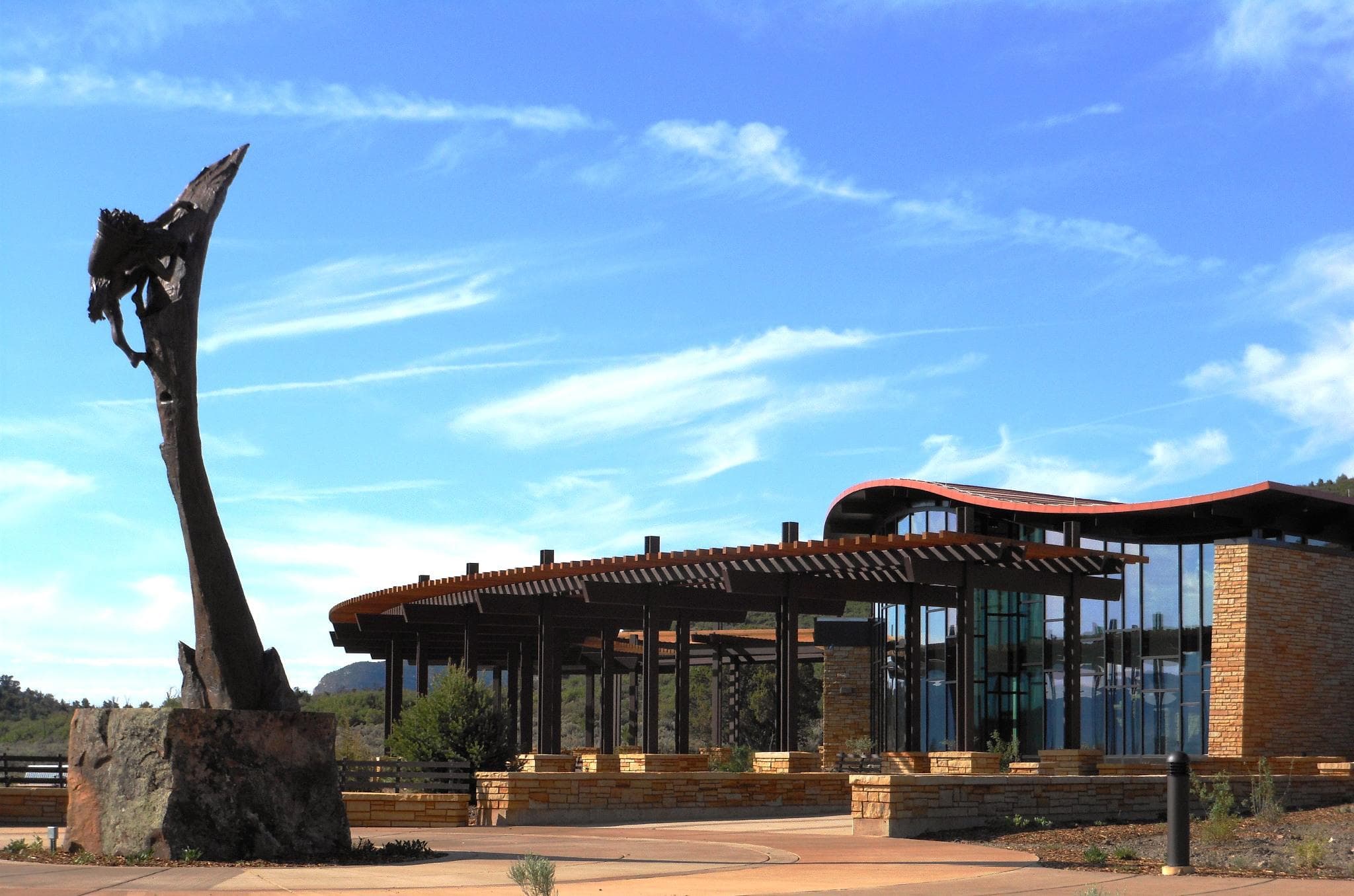 View of glass-fronted building with covered walkway and tall sculpture in front.