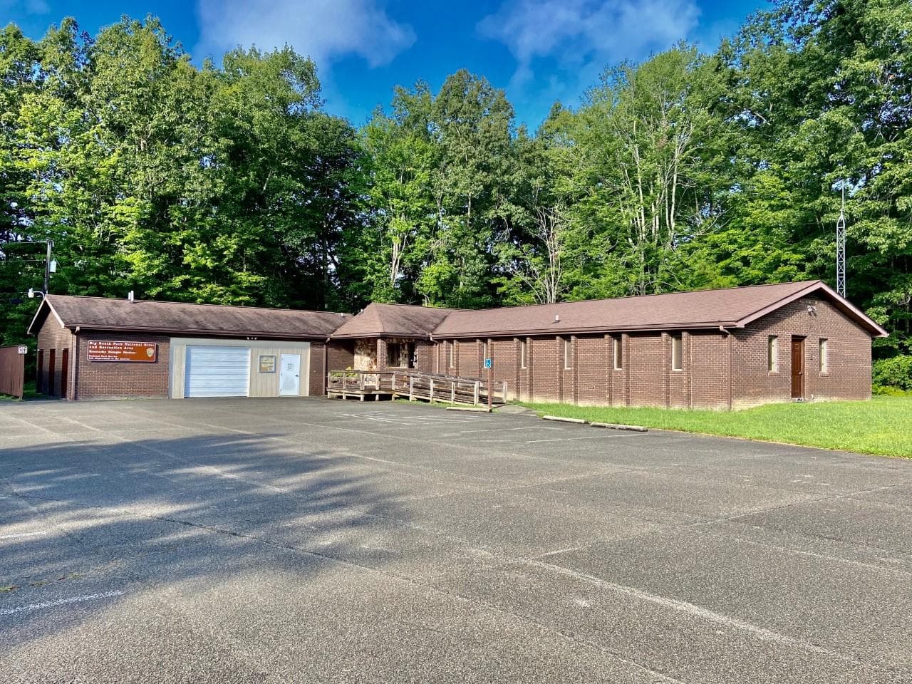 An L-shaped brown brick house with garage sits with a large paved parking lot in front.