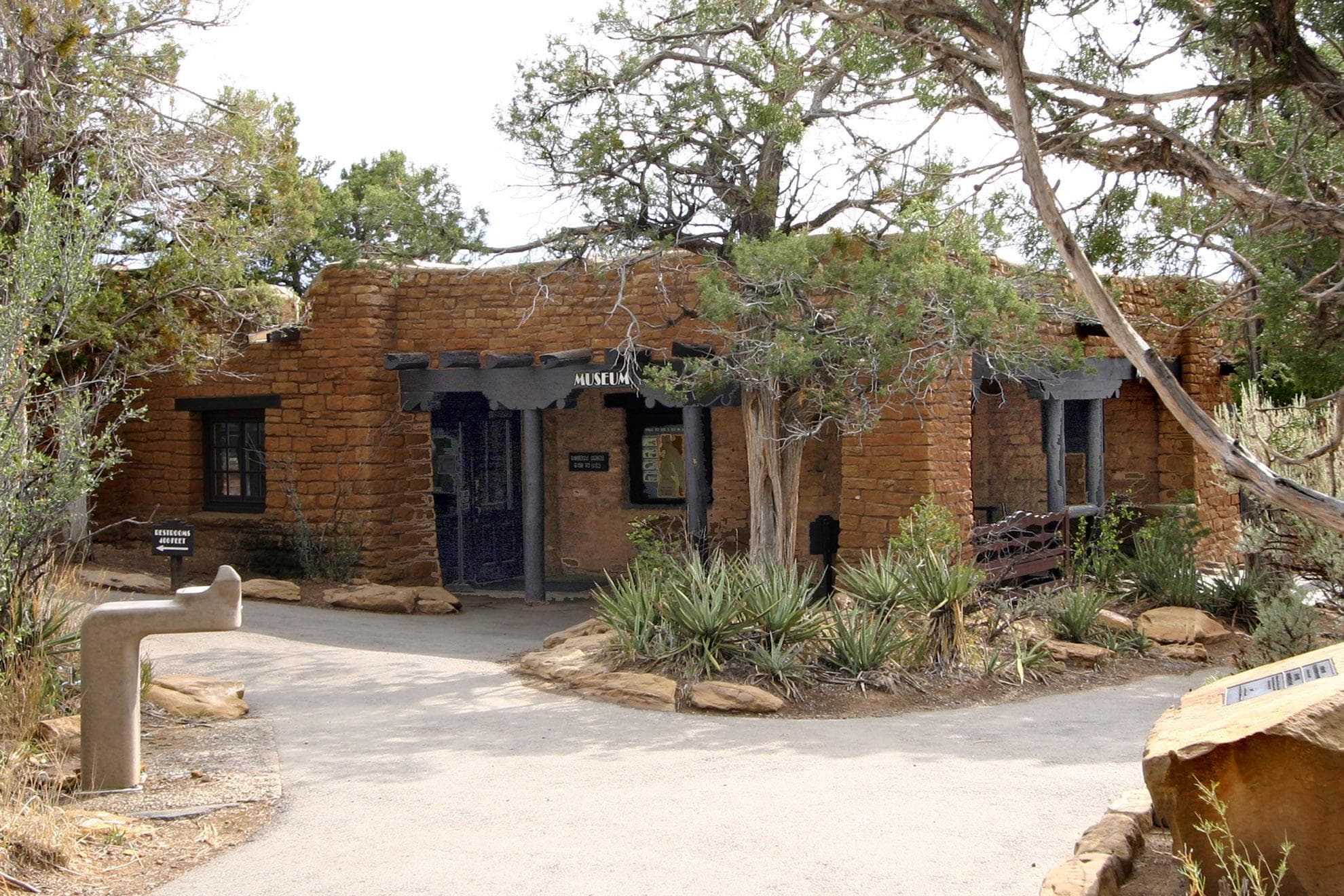 Stone-masonry building with covered entrance.Trees and plants surround it.