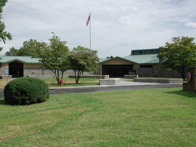 A gray stone building with green metal roof viewed across a grass field and road.