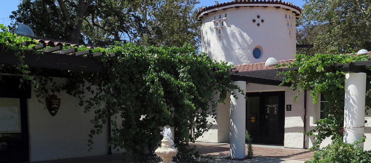 White building with brown roof with grape vines over walkways and fountain in front.