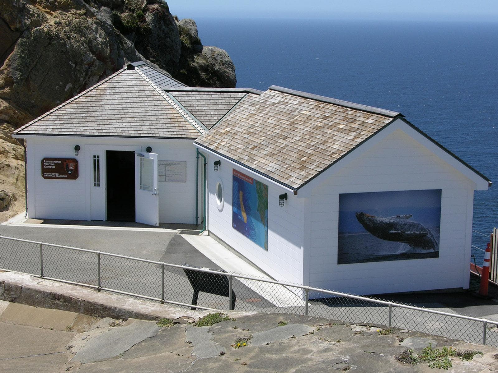 A smallish white-sided, brown-shingled building with two wings with the ocean in the background.