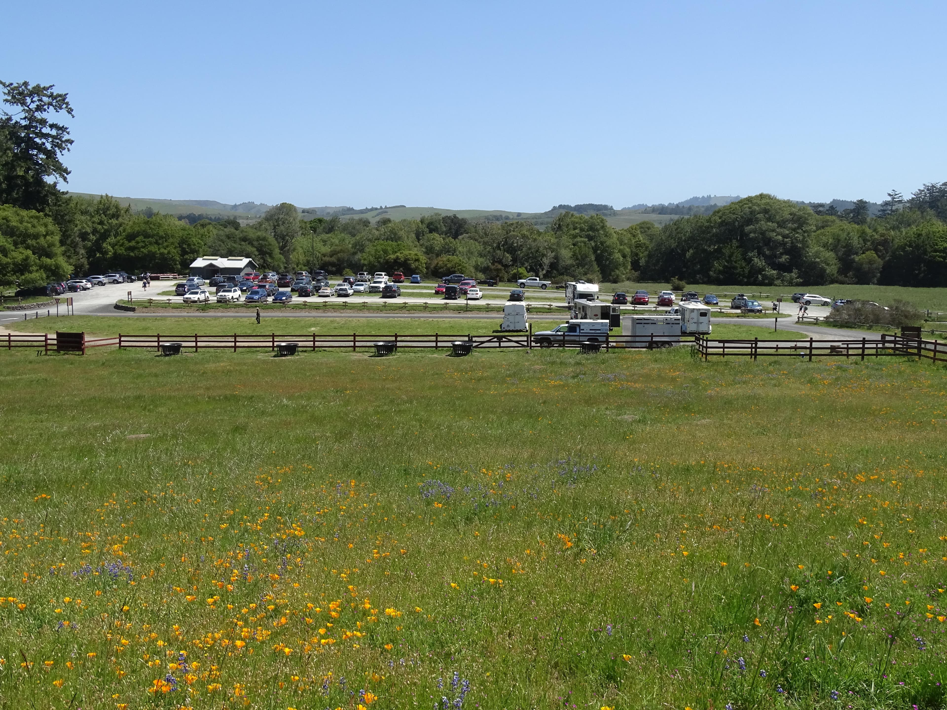 Looking across a grassy pasture to a large gravel parking lot partially filled with cars & trailers.