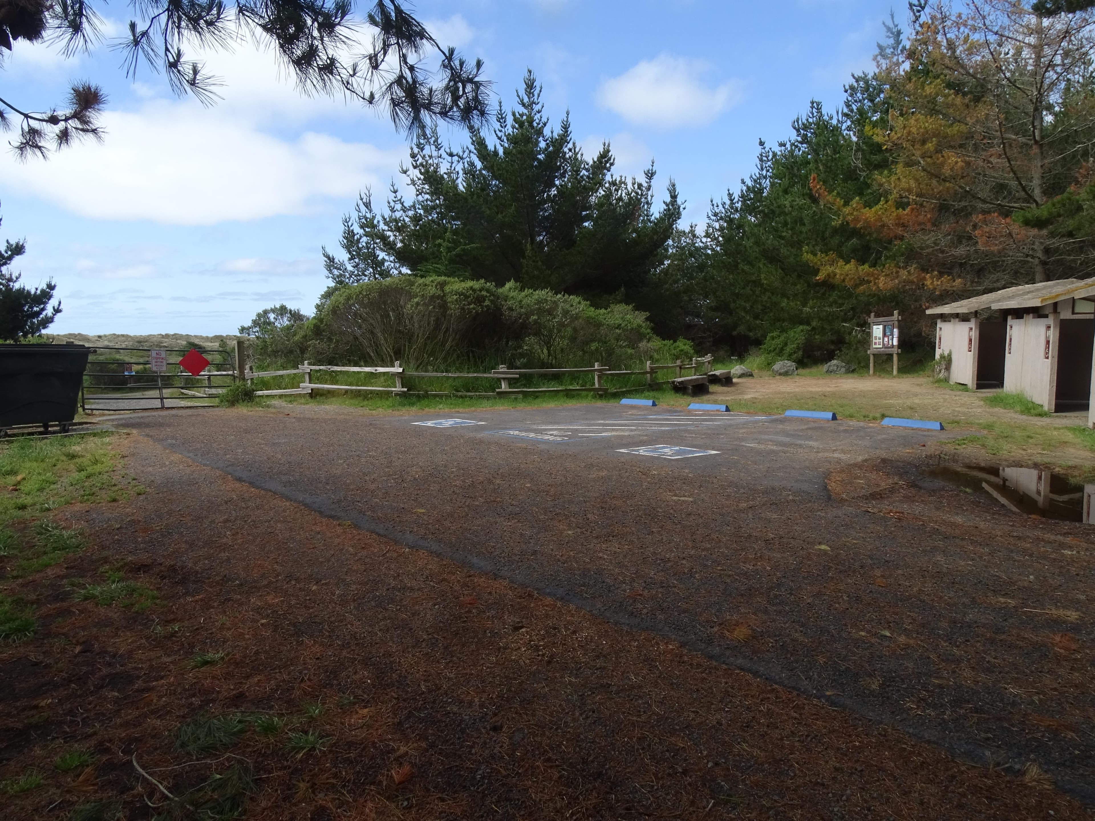 Two asphalt accessible parking spaces with vault toilets on the right & a fence & gate on the left.