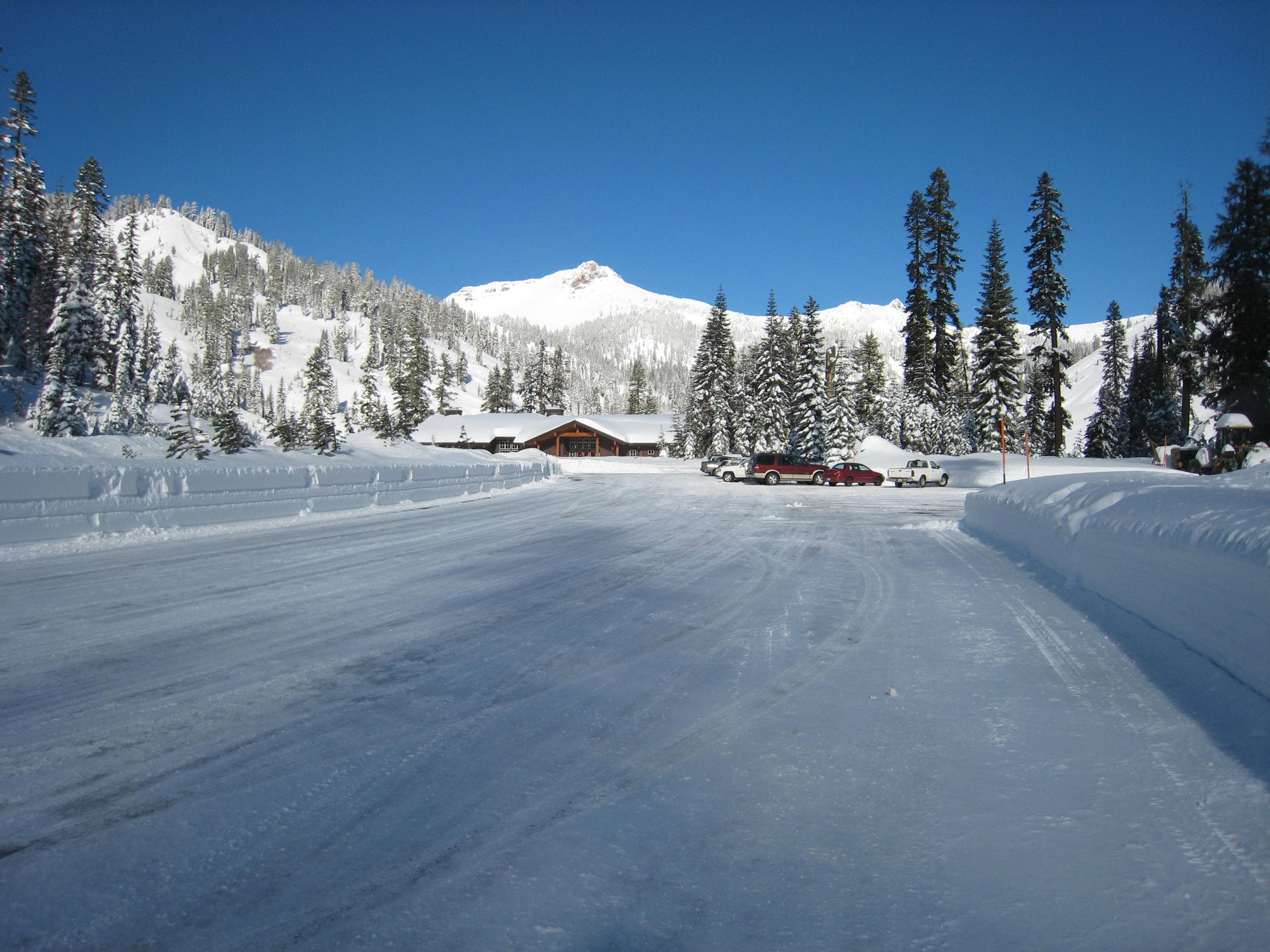 plowed parking lot with a few vehicles surrounded by deep snow, pine trees, and mountains