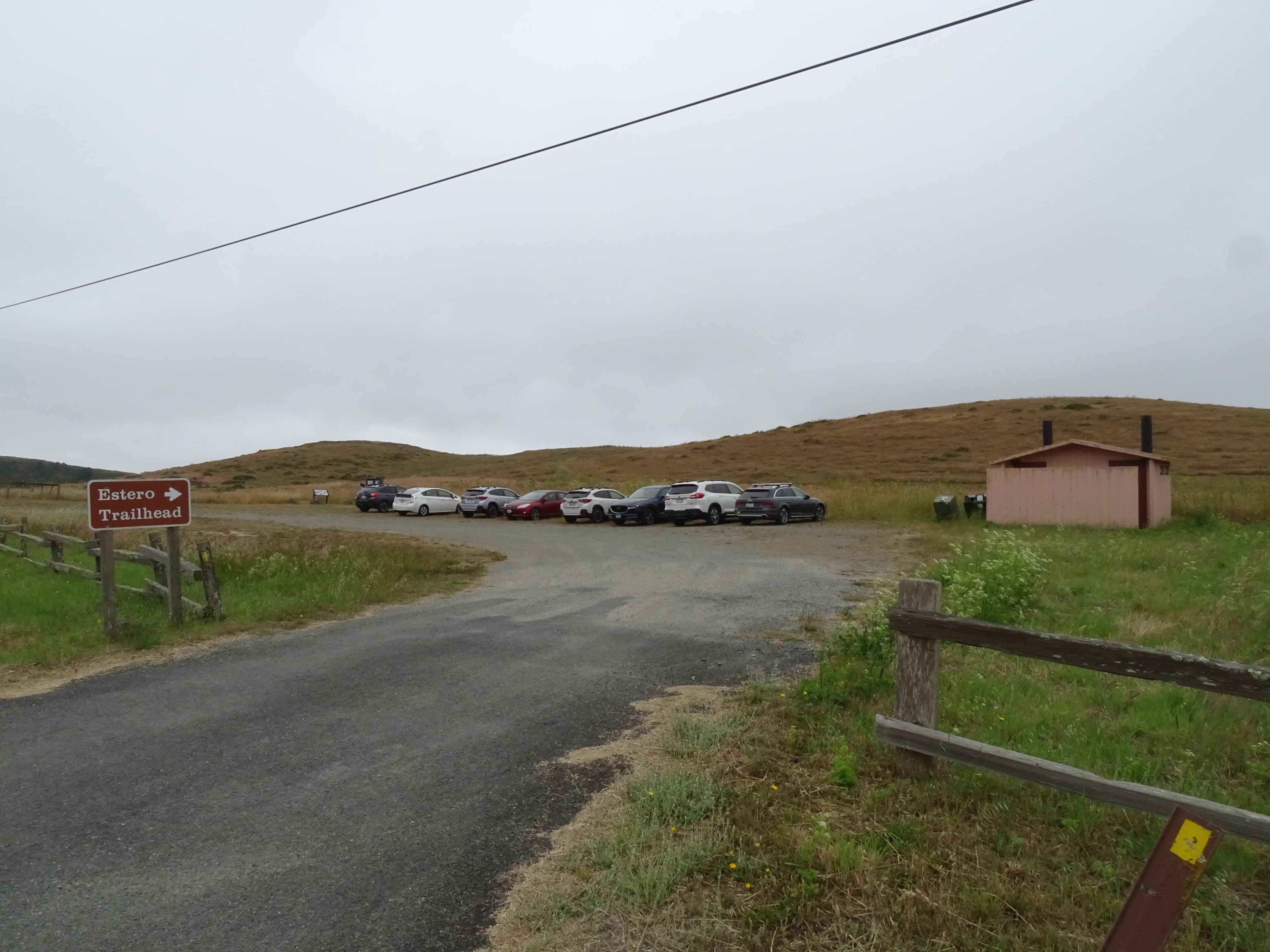 Eight vehicles in a small gravel parking lot with a vault toilet building on the right.