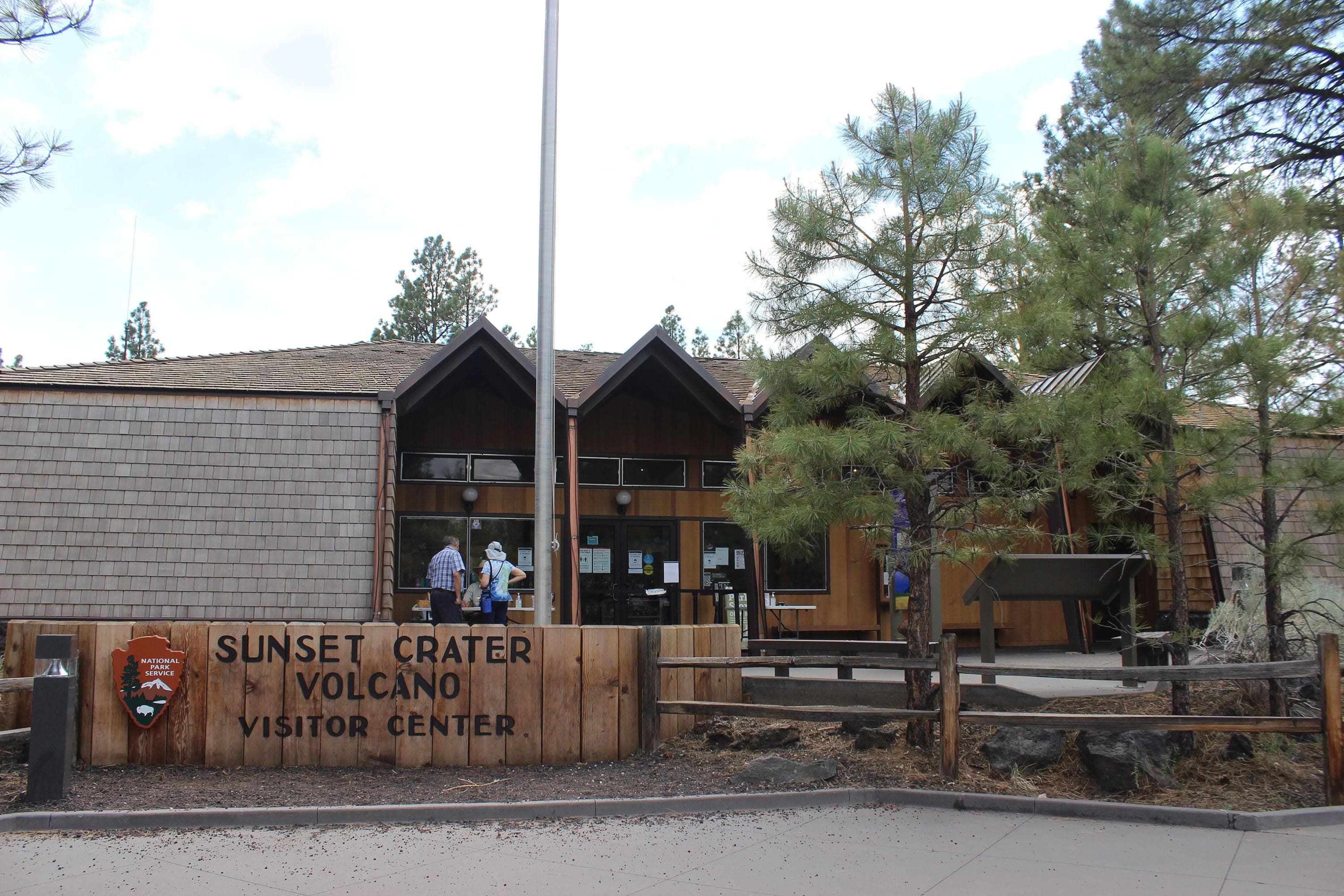 A wood and shingle building with a five gabled overhang, two trees, a flagpole, and an NPS park sign