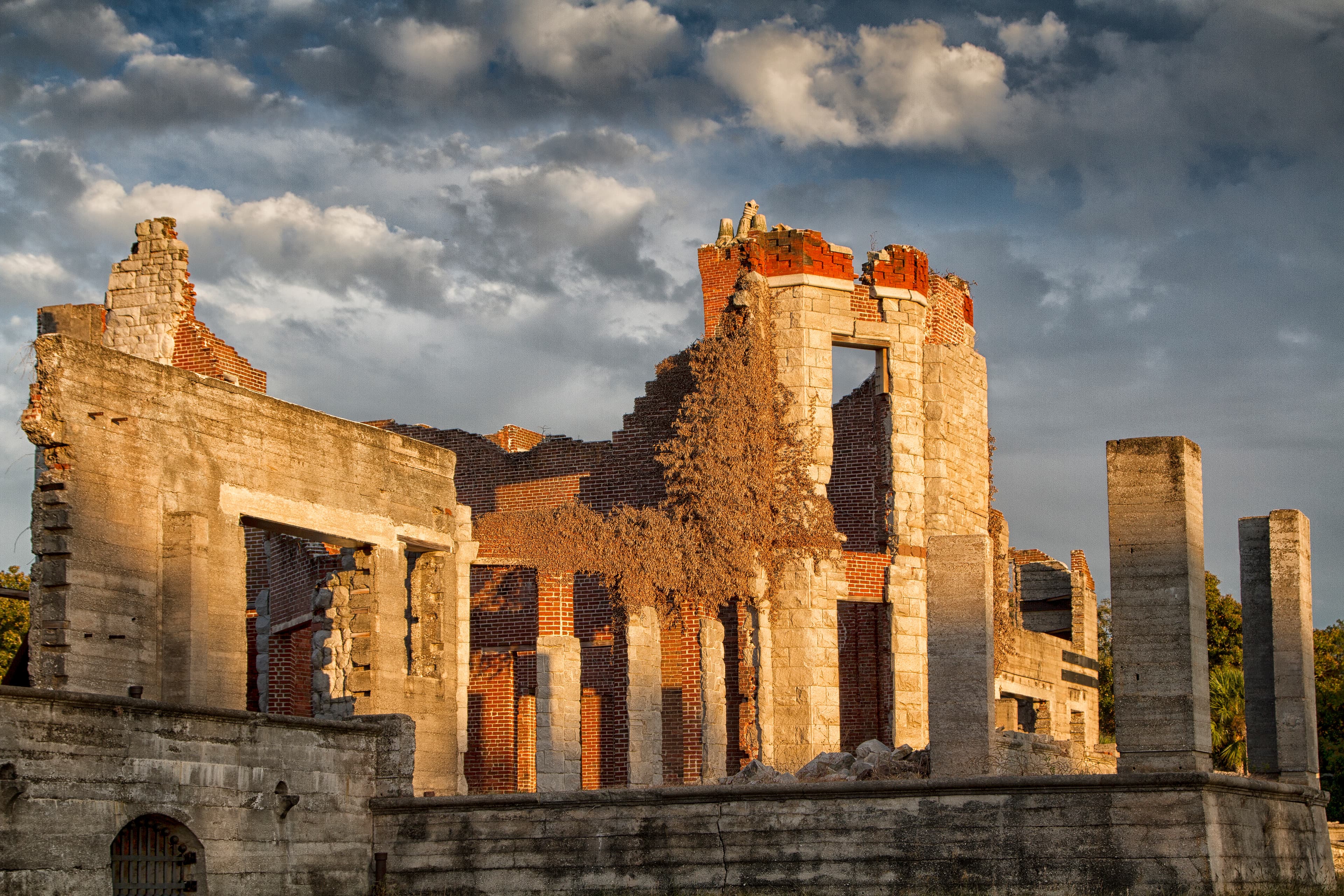 The brick and stonr ruins of a mansion are lit up by the evening sun with patchy clouds behind.