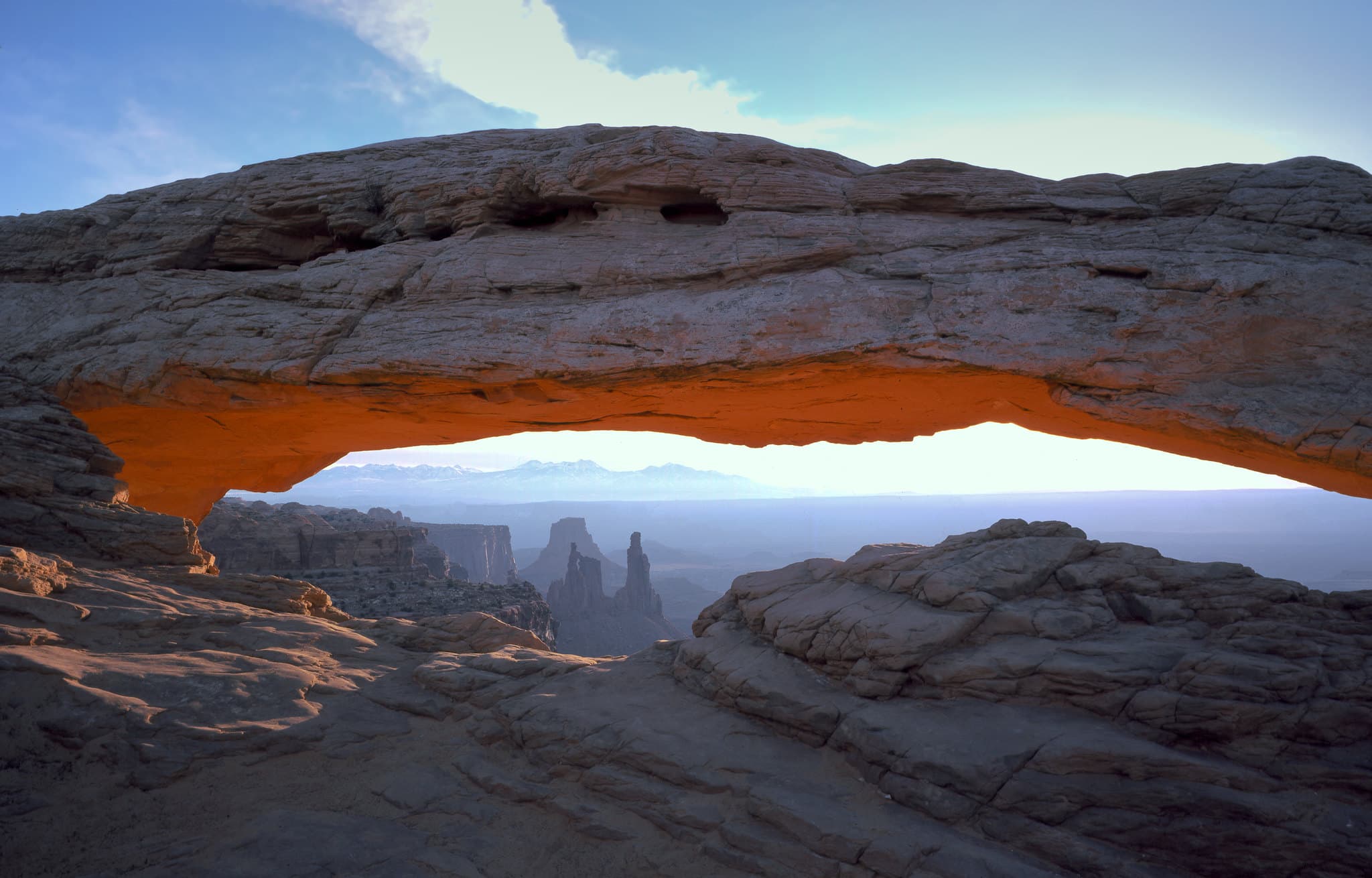 A thin arch is illuminated by sunrise, rock formations are visible in the distance.