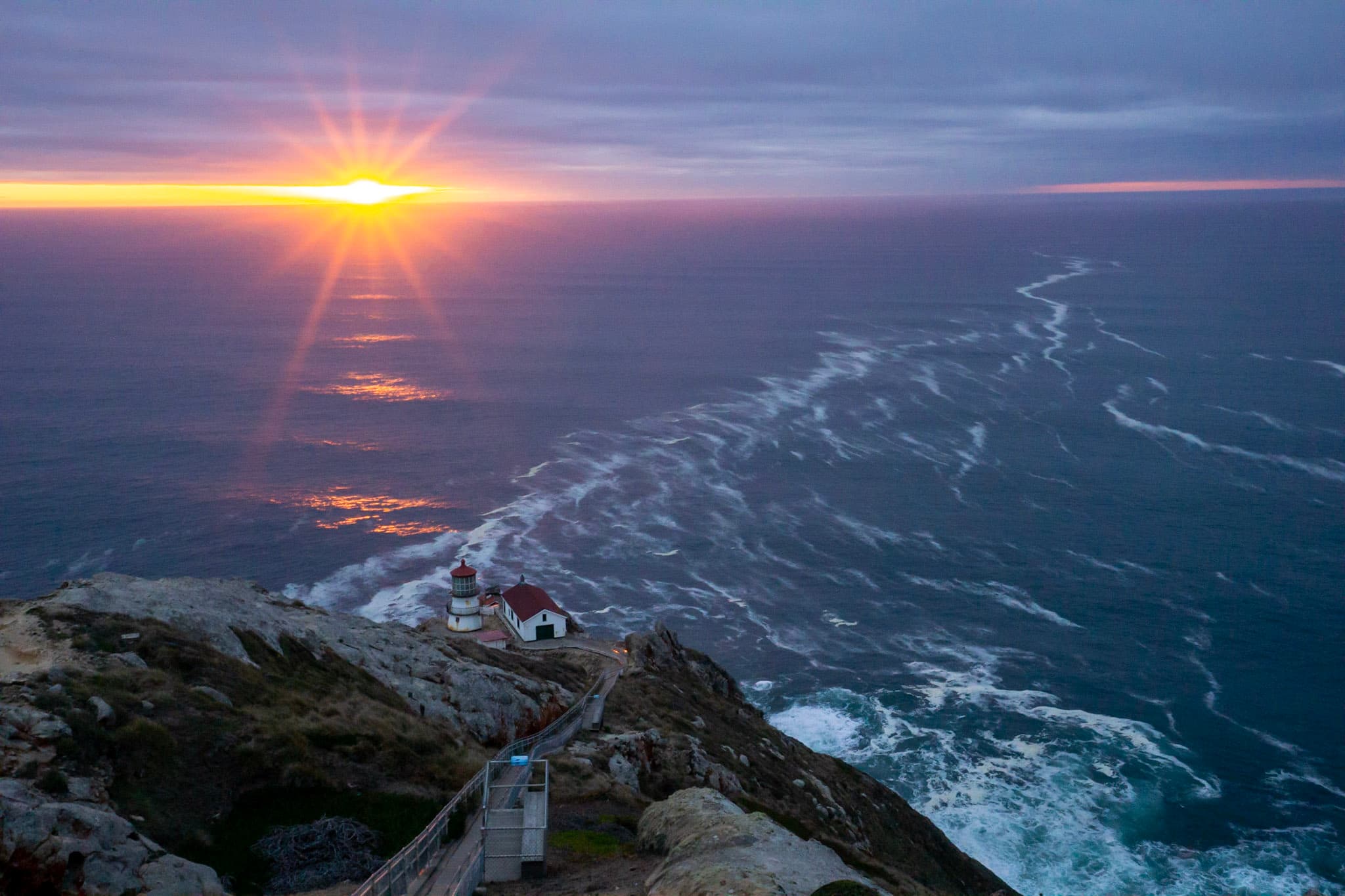 A short three story lighthouse on a rocky cliff overlooking the ocean at sunset.