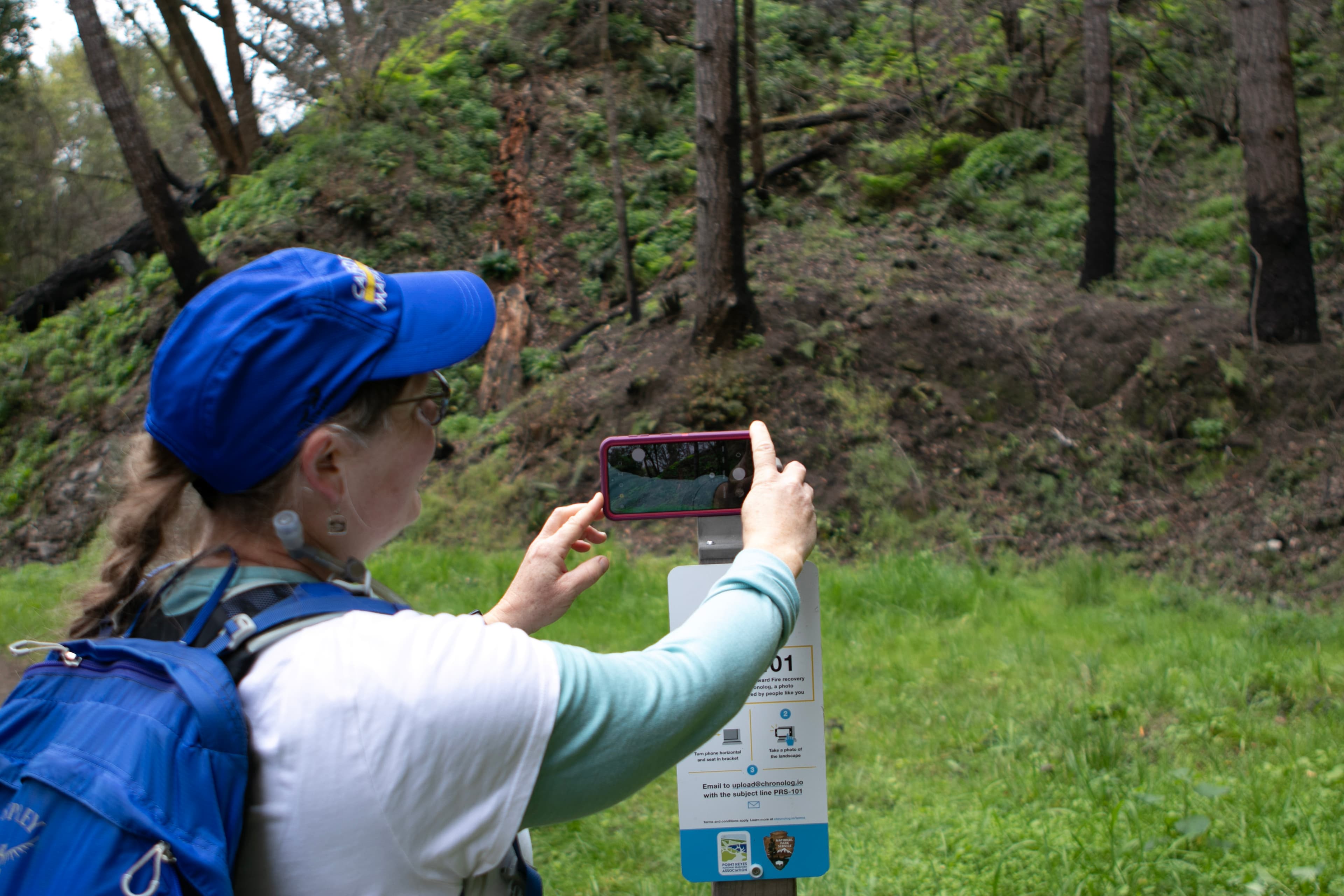 A woman takes a smartphone photo of a forested hill atop a bracket with a sign that reads "PRS 101"