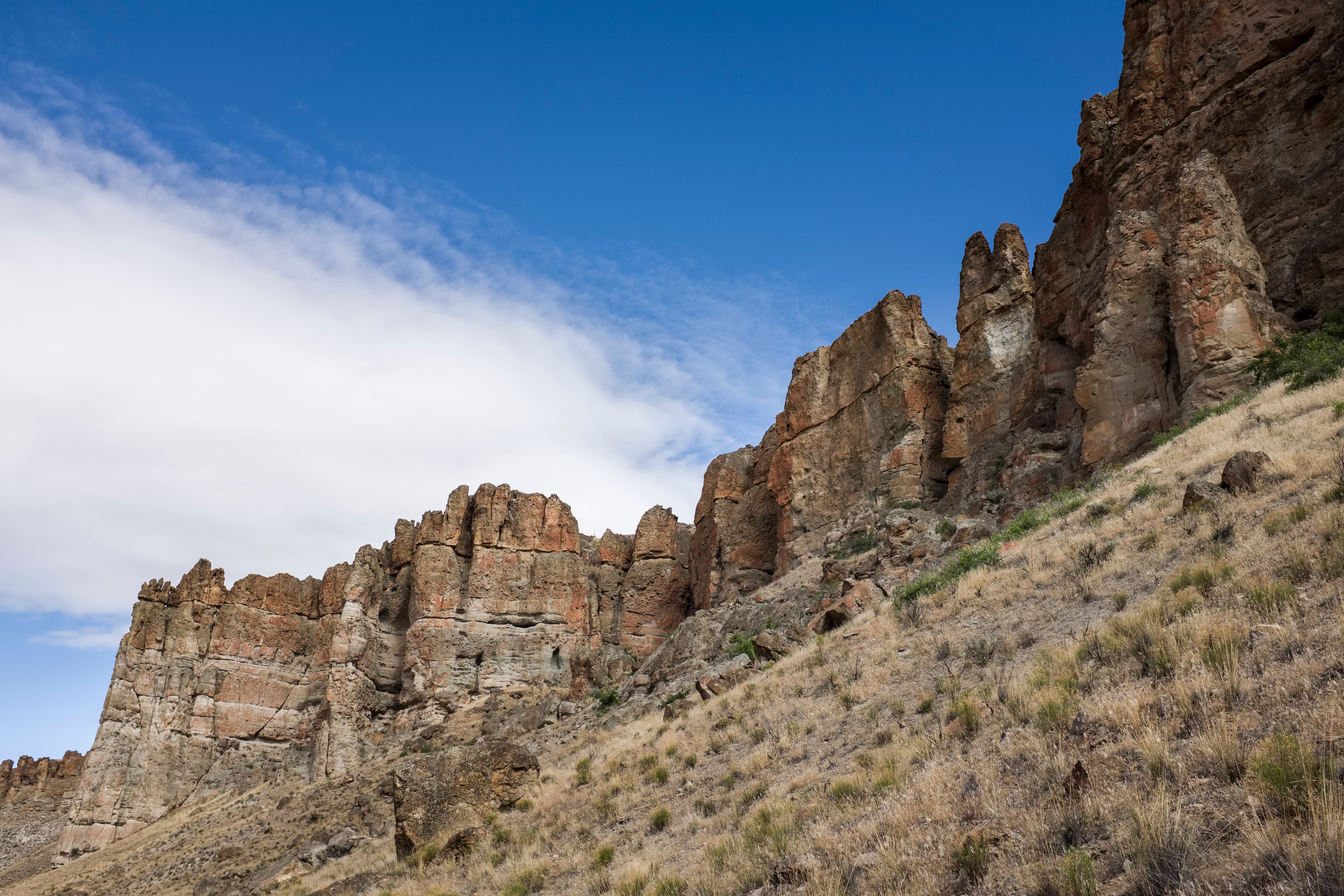Tan, towering mudflow cliff formations with grassy slopes at its base on a sunny day